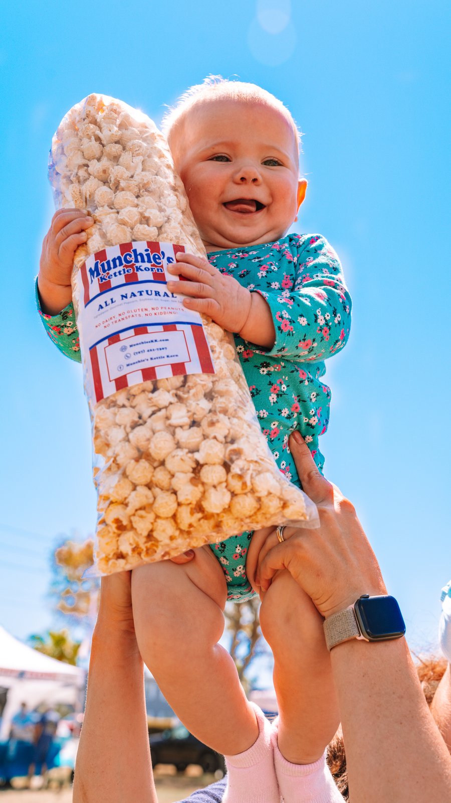 A baby in a floral outfit holding a large bag of Munchie's Kettle Korn popcorn outdoors under a blue sky.