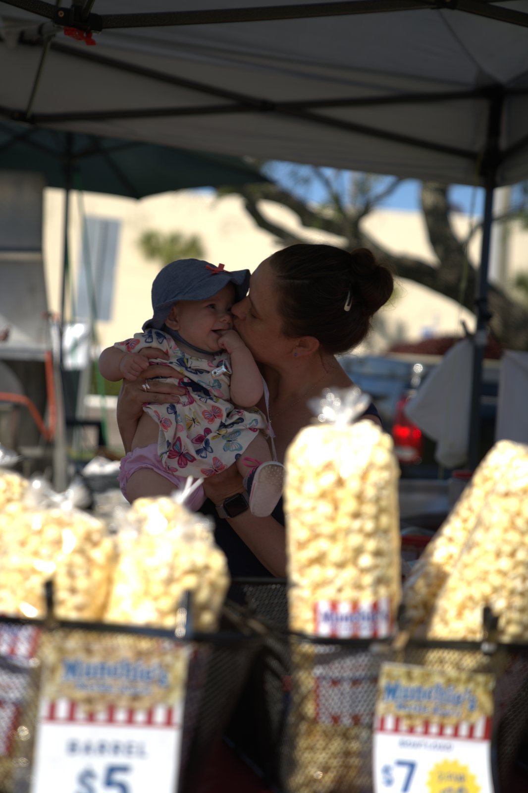 Kristin, founder of BoothBoss and Munchie's Kettle Korn, kissing her baby daughter RaeLynne at an outdoor kettle corn stand, bags of popcorn displayed in the foreground.
